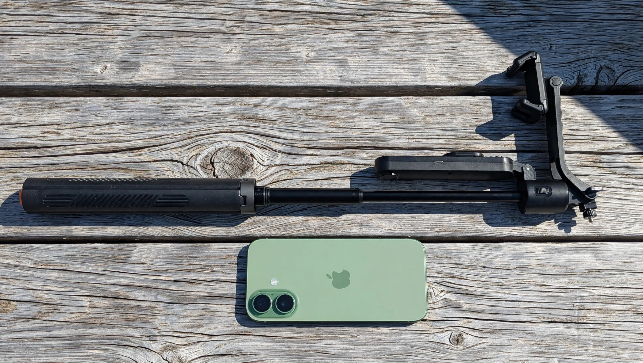 AI tracking selfie stick fully extended on weathered wood in harsh sunlight, green iPhone 17 laid below it